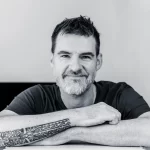 Guitarist Steve Allsworth standing with tattooed arms crossed in front of a computer
